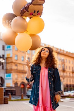 Genç, kahverengi kıvırcık saçlı, pembe uzun elbiseli, sarı, altın ve siyah balonlu bir kadın doğum günü partisine gitmek üzere sokakta yürüyor.