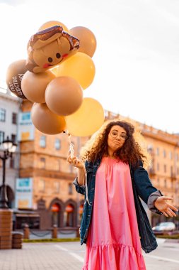 Genç, kahverengi kıvırcık saçlı, pembe uzun elbiseli, sarı, altın ve siyah balonlu bir kadın doğum günü partisine gitmek üzere sokakta yürüyor.
