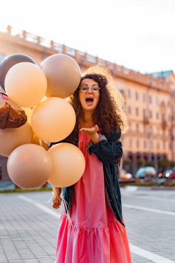Genç, kahverengi kıvırcık saçlı, pembe uzun elbiseli, sarı, altın ve siyah balonlu bir kadın doğum günü partisine gitmek üzere sokakta yürüyor.
