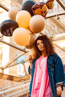 Genç, kahverengi kıvırcık saçlı, pembe uzun elbiseli, sarı, altın ve siyah balonlu bir kadın doğum günü partisine gitmek üzere sokakta yürüyor.
