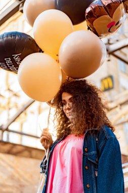 Genç, kahverengi kıvırcık saçlı, pembe uzun elbiseli, sarı, altın ve siyah balonlu bir kadın doğum günü partisine gitmek üzere sokakta yürüyor.