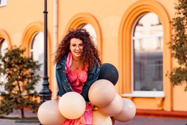 Genç, kahverengi kıvırcık saçlı, pembe uzun elbiseli, sarı, altın ve siyah balonlu bir kadın doğum günü partisine gitmek üzere sokakta yürüyor.