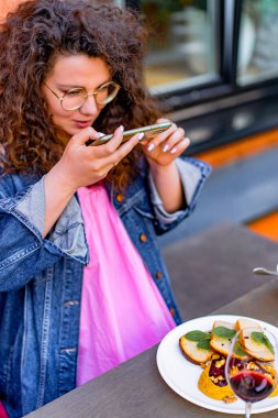 Kahverengi kıvırcık saçlı, bir kafede fotoğraf çeken güzel bir genç kadın telefonda yemek yiyor..