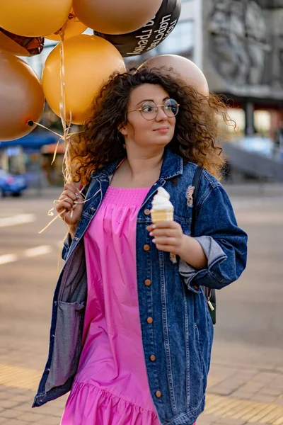 Pembe uzun elbiseli, sarı, altın ve siyah balonlu ve vanilyalı dondurma külahlı güzel, kahverengi kıvırcık saçlı kadın doğum günü partisine gitmek için caddede yürüyor.