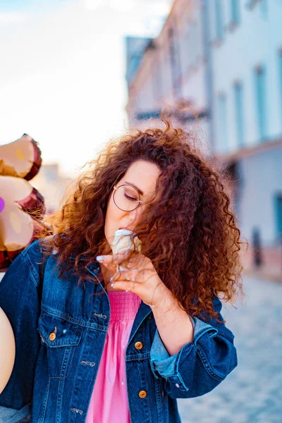 Pembe uzun elbiseli, sarı, altın ve siyah balonlu ve vanilyalı dondurma külahlı güzel, kahverengi kıvırcık saçlı kadın doğum günü partisine gitmek için caddede yürüyor.