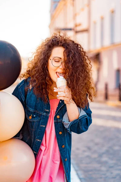 Pembe uzun elbiseli, sarı, altın ve siyah balonlu ve vanilyalı dondurma külahlı güzel, kahverengi kıvırcık saçlı kadın doğum günü partisine gitmek için caddede yürüyor.