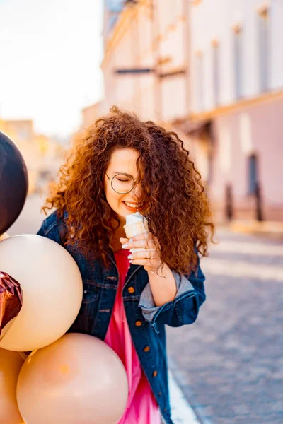 Pembe uzun elbiseli, sarı, altın ve siyah balonlu ve vanilyalı dondurma külahlı güzel, kahverengi kıvırcık saçlı kadın doğum günü partisine gitmek için caddede yürüyor.