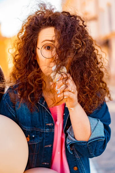 Pembe uzun elbiseli, sarı, altın ve siyah balonlu ve vanilyalı dondurma külahlı güzel, kahverengi kıvırcık saçlı kadın doğum günü partisine gitmek için caddede yürüyor.