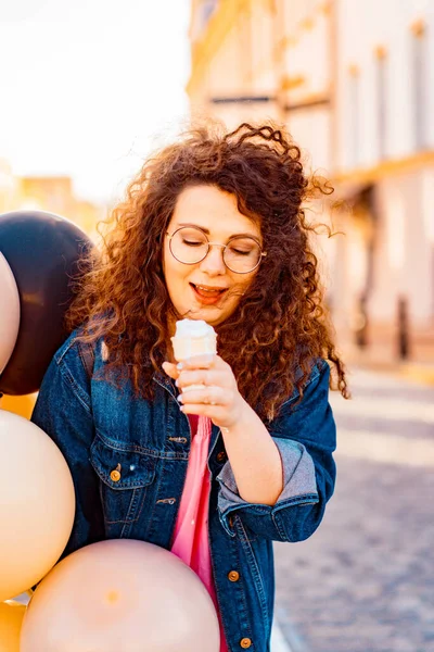 Pembe uzun elbiseli, sarı, altın ve siyah balonlu ve vanilyalı dondurma külahlı güzel, kahverengi kıvırcık saçlı kadın doğum günü partisine gitmek için caddede yürüyor.