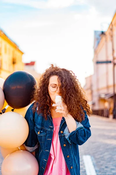 Pembe uzun elbiseli, sarı, altın ve siyah balonlu ve vanilyalı dondurma külahlı güzel, kahverengi kıvırcık saçlı kadın doğum günü partisine gitmek için caddede yürüyor.