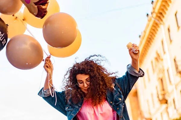 Pembe uzun elbiseli, sarı, altın ve siyah balonlu ve vanilyalı dondurma külahlı güzel, kahverengi kıvırcık saçlı kadın doğum günü partisine gitmek için caddede yürüyor.