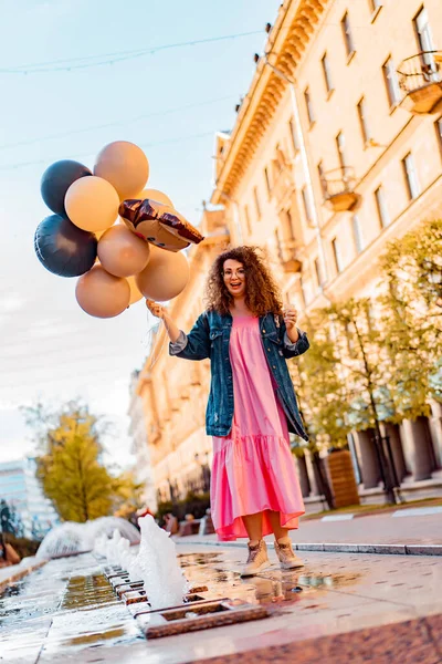 Pembe uzun elbiseli, sarı, altın ve siyah balonlu ve vanilyalı dondurma külahlı güzel, kahverengi kıvırcık saçlı kadın doğum günü partisine gitmek için caddede yürüyor.