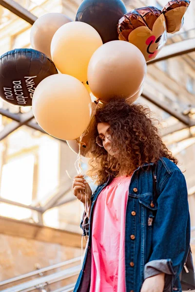Genç, kahverengi kıvırcık saçlı, pembe uzun elbiseli, sarı, altın ve siyah balonlu bir kadın doğum günü partisine gitmek üzere sokakta yürüyor.