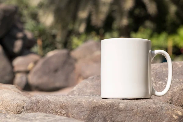 白色空白咖啡杯模仿 特写的杯子外面在阳光下一些大型装饰花园石头 完美的企业销售杯子 只是覆盖你的报价或设计的图像 — 图库照片