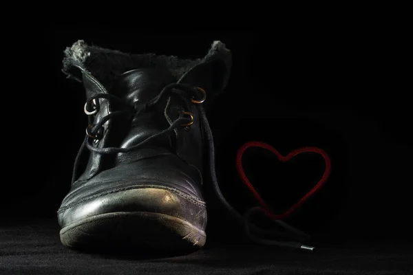 Old black shoe forms a red heart with its shoelace against a dark background, concept for love in old age or greeting card for valentine's day — Stock Photo, Image