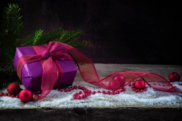 Weihnachts- und Neujahrsgeschenk aus violettem Papier, Bändern und Kugeln auf rustikalem Holzbrett mit Schnee, dunklem Hintergrund — Stockfoto