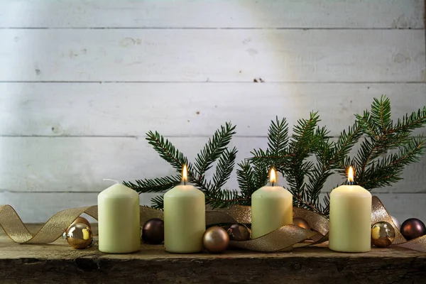 Três velas brancas acesas no terceiro advento, decoração de Natal com galhos de abeto, bugigangas e uma fita dourada em uma tábua de madeira rústica contra a madeira branca — Fotografia de Stock