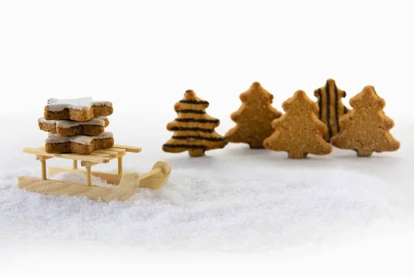 Wooden sled carries cinnamon star cookies through the snow in front of christmas trees made from gingerbread — Stock Photo, Image