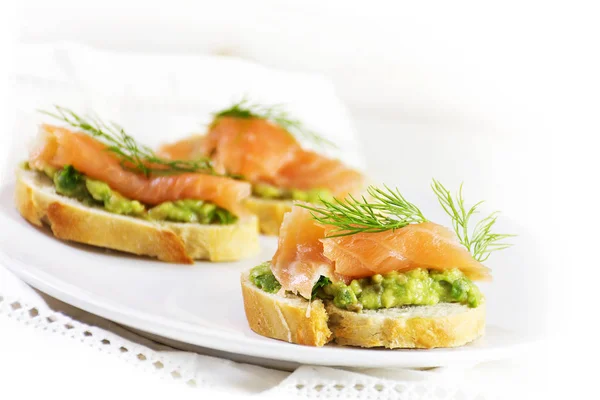 Smoked salmon and avocado cream or guacamole on baguette slices  on a white plate as a healthy party snack, bright background copy space — Stock Photo, Image