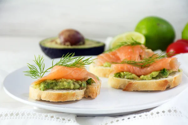 Sandwiches de baguette con salmón ahumado y crema de aguacate o guacamole como canapés de fiesta saludables en un plato blanco, ingredientes en el fondo brillante — Foto de Stock