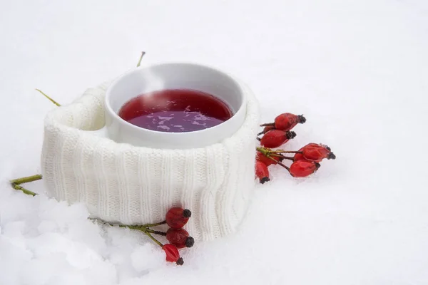 Té rojo caliente en la nieve, taza con bufanda y rosa mosqueta contra el resfriado común, fondo blanco nevado con espacio para copiar — Foto de Stock