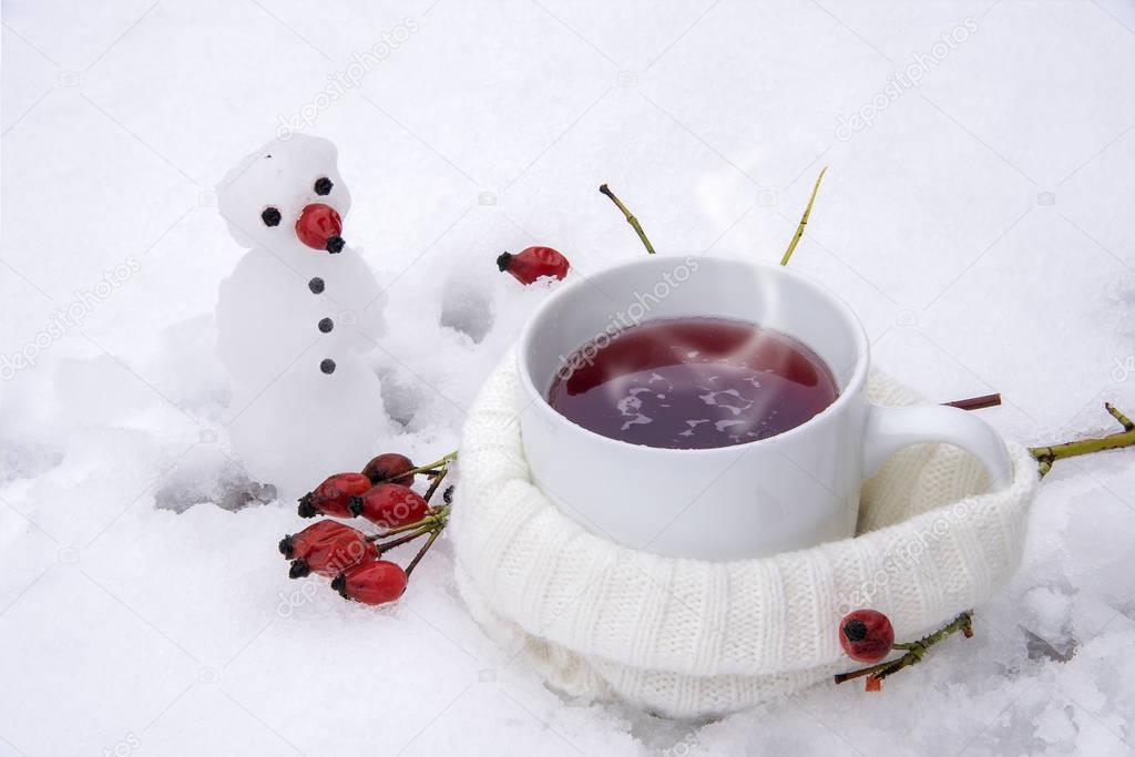 Hot tea with red rose hips  in the snow and a little snowman, winter drink against influenza