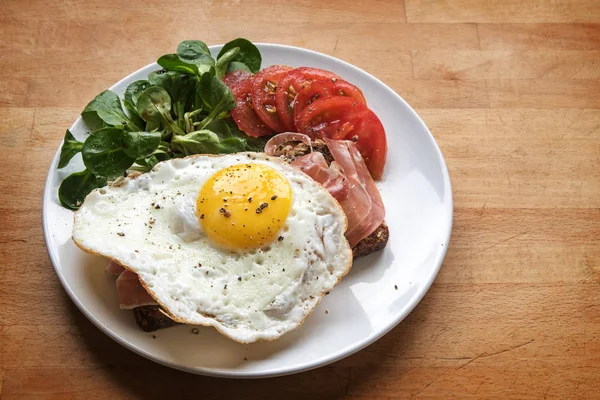 Huevo frito sobre pan con jamón adornado con ensalada verde y tomates en un plato blanco sobre una mesa de madera, espacio para copiar —  Fotos de Stock