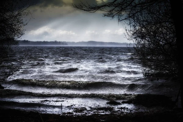 Jezero s tmavě vlnami na břeh v špatné bouřlivé počasí v lese a sluneční paprsky na obloze, na šířku — Stock fotografie