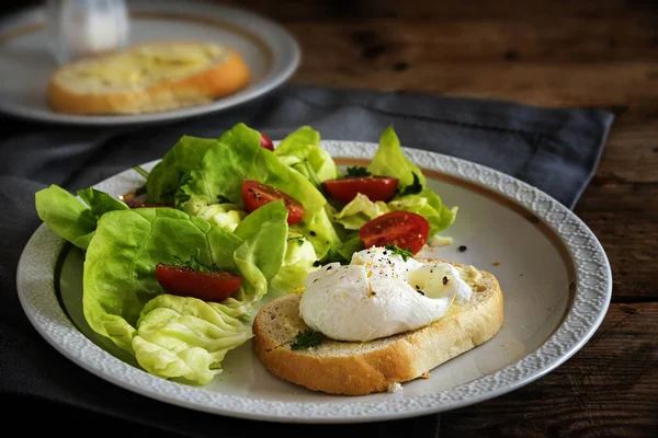 Huevo escalfado en rebanada de baguette con ensalada y tomates, plato blanco sobre una mesa de madera rústica oscura — Foto de Stock