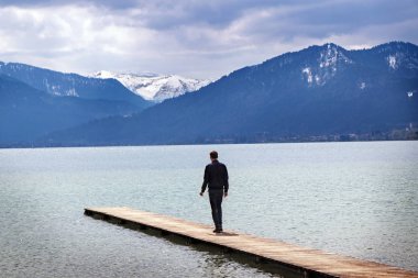 Genç adam tek başına tegernsee Gölü'iskele üzerinde durur ve mavi bakar kar kaplı dağlar ünlü turistik beldesinde Bavyera Alpleri