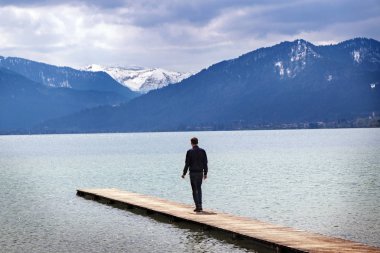 Young man stands alone on the jetty in the tegernsee lake and looks at the blue snow covered mountains in the famous tourist resort of the Bavarian Alps clipart