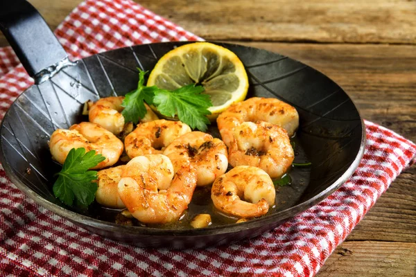 Fried tiger shrimps with garlic, lemon and italian parsley garnish in a black iron pan on a napkin and a rustic wooden table — Stock Photo, Image