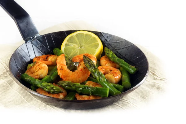 Fried prawns or shrimps with  green asparagus peaks and a lemon slice in a black iron pan, corner background fades to white — Stock Photo, Image