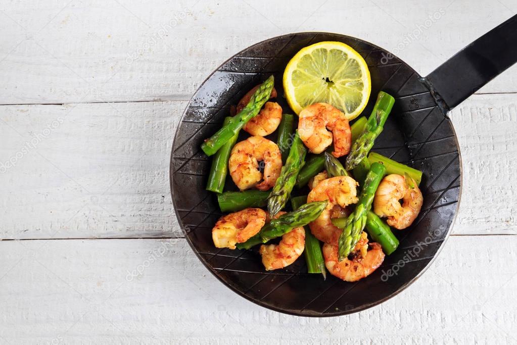 fried prawns or shrimps with  green asparagus peaks and a lemon slice in a black iron pan on rustic white painted wood with copy space, top view
