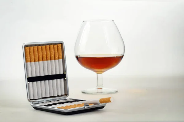 Табак и алкоголь, портсигар с фильтрующими сигаретами и стакан с коньяком или бренди на светло-сером фоне с копировальным пространством — стоковое фото