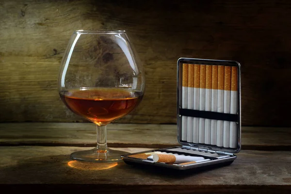 Alcohol and tobacco, glass with cognac or brandy and a case with cigarettes on warm rustic wood, concept for enjoyment or addiction — Stock Photo, Image