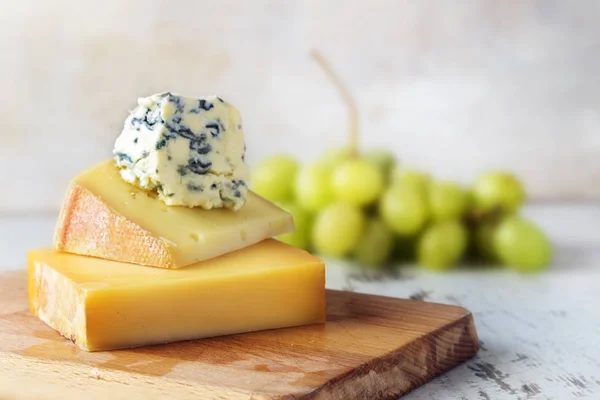 Pile de différents fromages sur une planche de bois, raisins verts flous dans le fond rustique lumineux — Photo