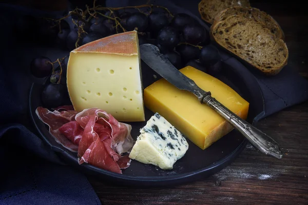 Supper plate with various cheese, air dried ham, blue grapes and bread on dark rustic wood — Stock Photo, Image