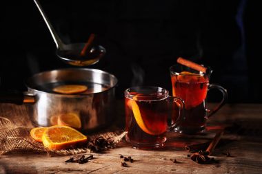 bir tencerede pişirilmiş sıcak mulled şarap buğulaması ve cam bardağı portakal dilimleri, karanfil, yıldız anason ve karanlık bir arka plan karşı rustik ahşap bir masa üzerine tarçın gibi Noel baharatlar ile servis
