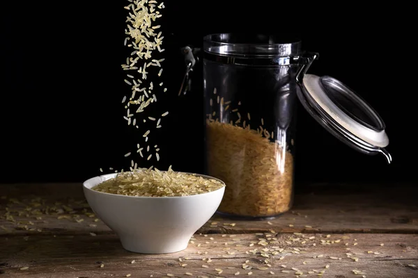 Granos de arroz cayendo en un tazón blanco junto a un frasco de vidrio con arroz sobre una mesa de madera rústica sobre un fondo oscuro — Foto de Stock