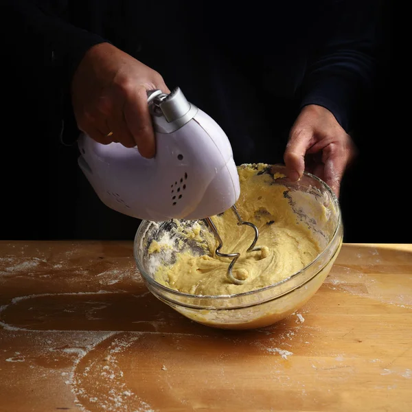 Женские руки перемешивают тесто со смесителем для торта или хлеба в миске на деревянной кухонной столешнице, подготовка к выпечке, темный фон с копировальным пространством — стоковое фото