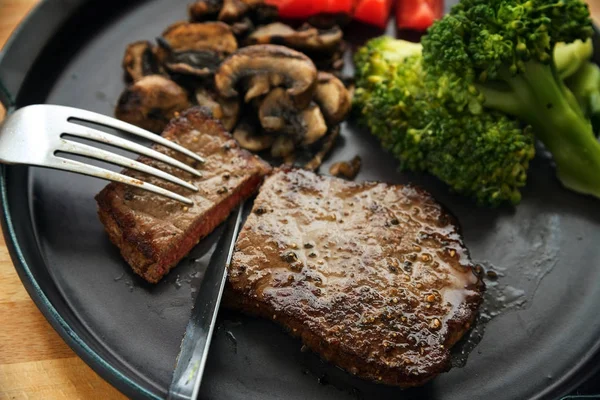 Beef steak is cut with knife and fork, with vegetables like broccoli, mushrooms and tomatoes, low carb diet dinner on a dark gray plate — Stock Photo, Image