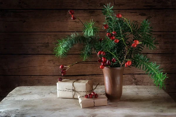 Tannenzweige und Hagebutten in einer Keramikvase und Weihnachtsgeschenkschachteln auf einem rustikalen Tisch vor einer dunklen Holzwand mit Kopierraum, natürlicher ländlicher Herbst- und Winterdekoration — Stockfoto