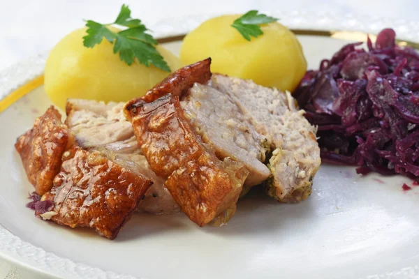 Svinekjøtt med sprø skorpe, med poteter, persille og rødkål på en hvit tallerken, som nærmer seg – stockfoto