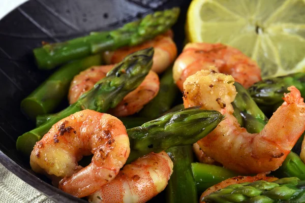 Fried prawns or shrimps with  green asparagus peaks and a lemon slice in a black iron pan, close up — Stock Photo, Image