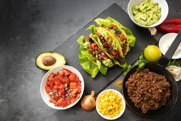 Tacos mexicanos e ingredientes como carne frita moída, salsa de tomate, guacamole, milho e especiarias em um prato de ardósia escuro com espaço de cópia — Fotografia de Stock