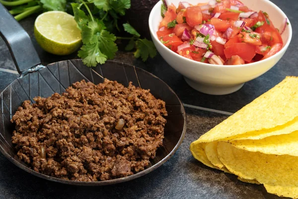 Sült darált marhahús, egy serpenyőben, mint a töltés taco kagyló alapanyagokból, paradicsom salsa-, mész- és gyógynövények a sötét kő háttér — Stock Fotó