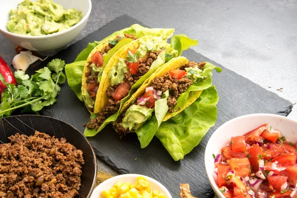 Mexican tacos and some ingredients like fried ground beef, tomato salsa, guacamole, corn and herbs on a dark slate plate with copy space — Stock Photo, Image