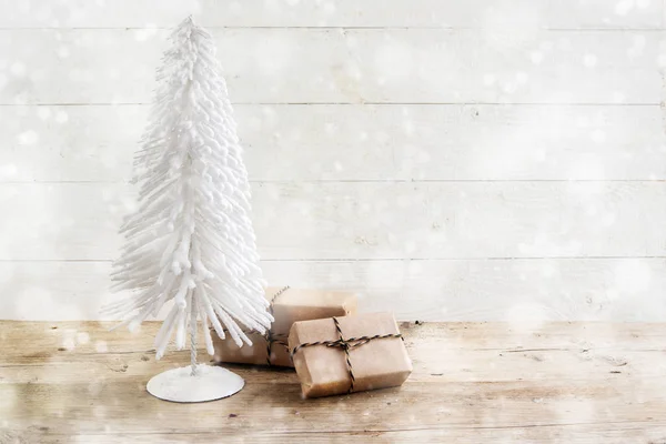 Kleiner Weihnachtsbaum aus weißem Draht und Geschenken in Kraftpapier auf rustikalem Holztisch, weiß lackierter Vintage-Hintergrund mit schneebedeckten Bokeh-Lichtern, Kopierraum — Stockfoto