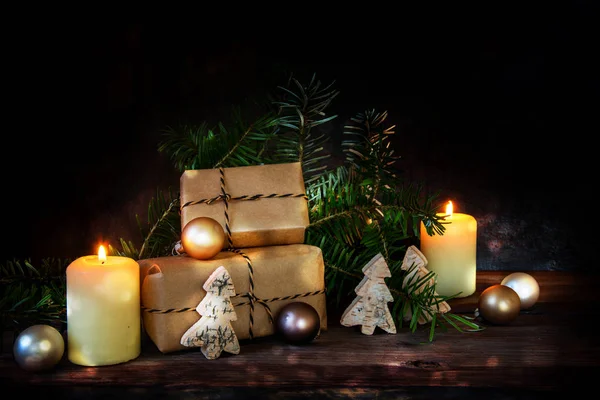 Caixas de presente em papel kraft e duas velas em chamas decoradas com bolas de Natal, ramos de abeto e pequenas árvores de madeira em um fundo rústico escuro com espaço de cópia — Fotografia de Stock